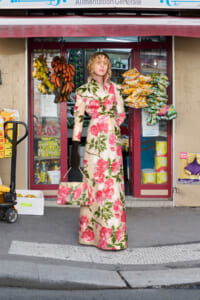 Person posing in a long cream coat with large pink flower print and black gloves, standing in front of a small shop with produce bags hanging outside.