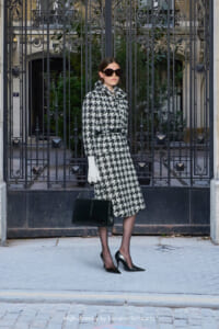 Woman in a black-and-white houndstooth coat with a belt, white gloves, oversized sunglasses, and a black handbag posing before an ornate iron gate.