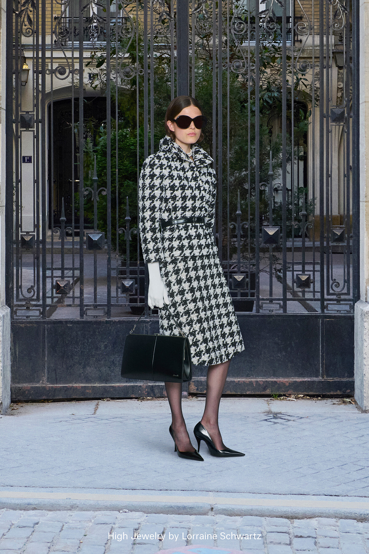 Woman in a black-and-white houndstooth coat with a belt, white gloves, oversized sunglasses, and a black handbag posing before an ornate iron gate.