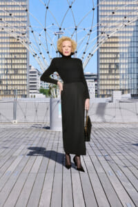 Elegant woman in a black turtleneck dress posing on a wooden deck with a geometric metal arch sculpture behind her.