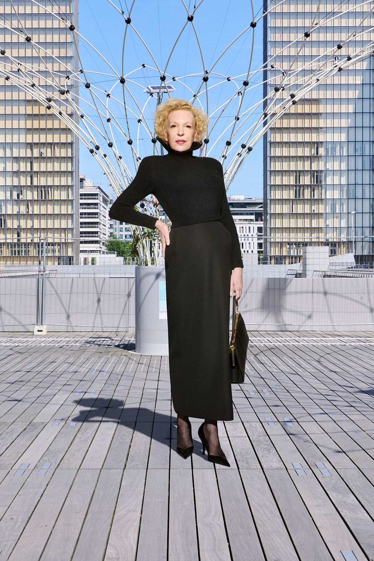 Elegant woman in a black turtleneck dress posing on a wooden deck with a geometric metal arch sculpture behind her.
