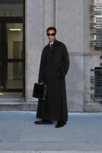 Man in a long black overcoat and sunglasses stands outside a building, holding a black briefcase.