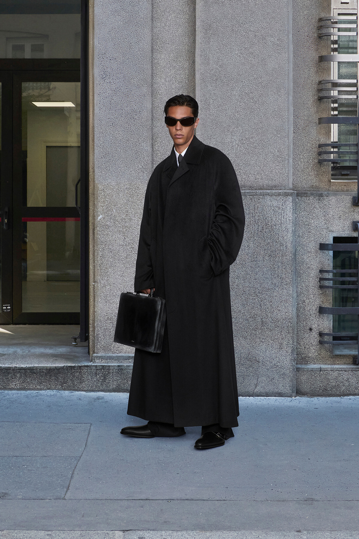 Man in a long black overcoat and sunglasses stands outside a building, holding a black briefcase.