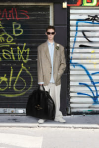 Man in oversized beige suit with light blue shirt and white sneakers, holding a large black duffel bag, standing against graffiti-covered shutters wearing dark sunglasses.
