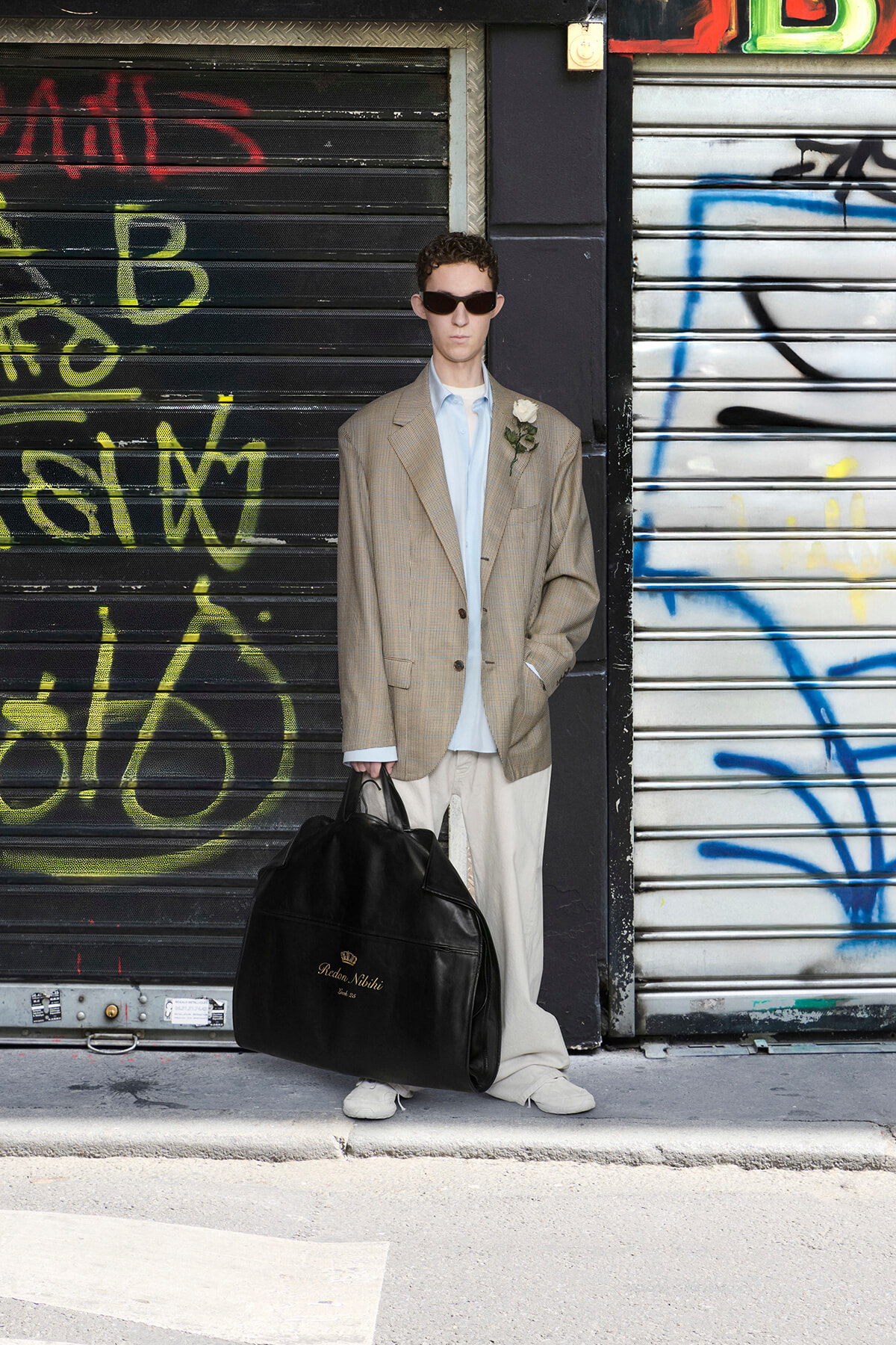 Man in oversized beige suit with light blue shirt and white sneakers, holding a large black duffel bag, standing against graffiti-covered shutters wearing dark sunglasses.