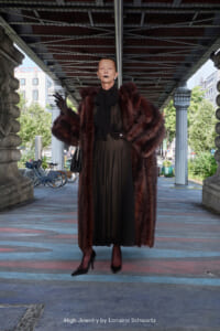 Older woman in a black dress and burgundy fur coat stands under an urban bridge, wearing gloves and carrying a small black handbag.