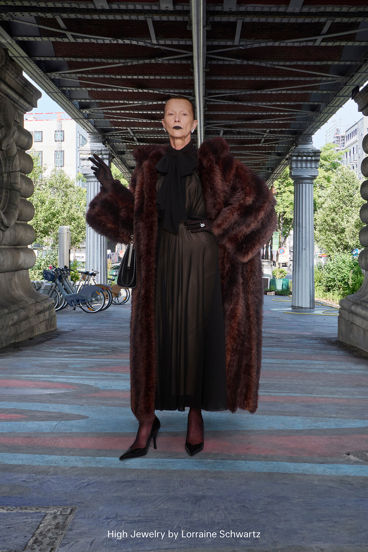 Older woman in a black dress and burgundy fur coat stands under an urban bridge, wearing gloves and carrying a small black handbag.
