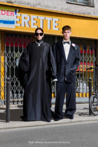 Two fashion models pose on a sidewalk in front of a bright yellow storefront; woman in a black gown with oversized sunglasses and jewelry, man in a black tuxedo with a boutonniere.
