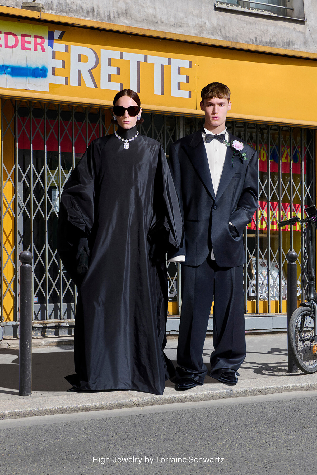 Two fashion models pose on a sidewalk in front of a bright yellow storefront; woman in a black gown with oversized sunglasses and jewelry, man in a black tuxedo with a boutonniere.