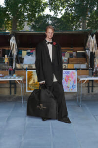 Man in a black tuxedo with a red rose boutonniere stands in front of a market stall with paintings, holding a large black garment bag.