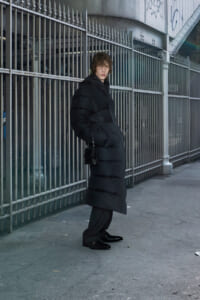 Young man wearing a long black puffer coat stands against a tall metal fence on an urban street.