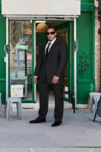 Man in a black suit and sunglasses standing in the entrance of a green storefront.