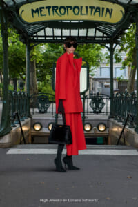 Model in a bright red coat and matching pants with black gloves and handbag posing at a Paris Métropolitain entrance, wearing sunglasses.