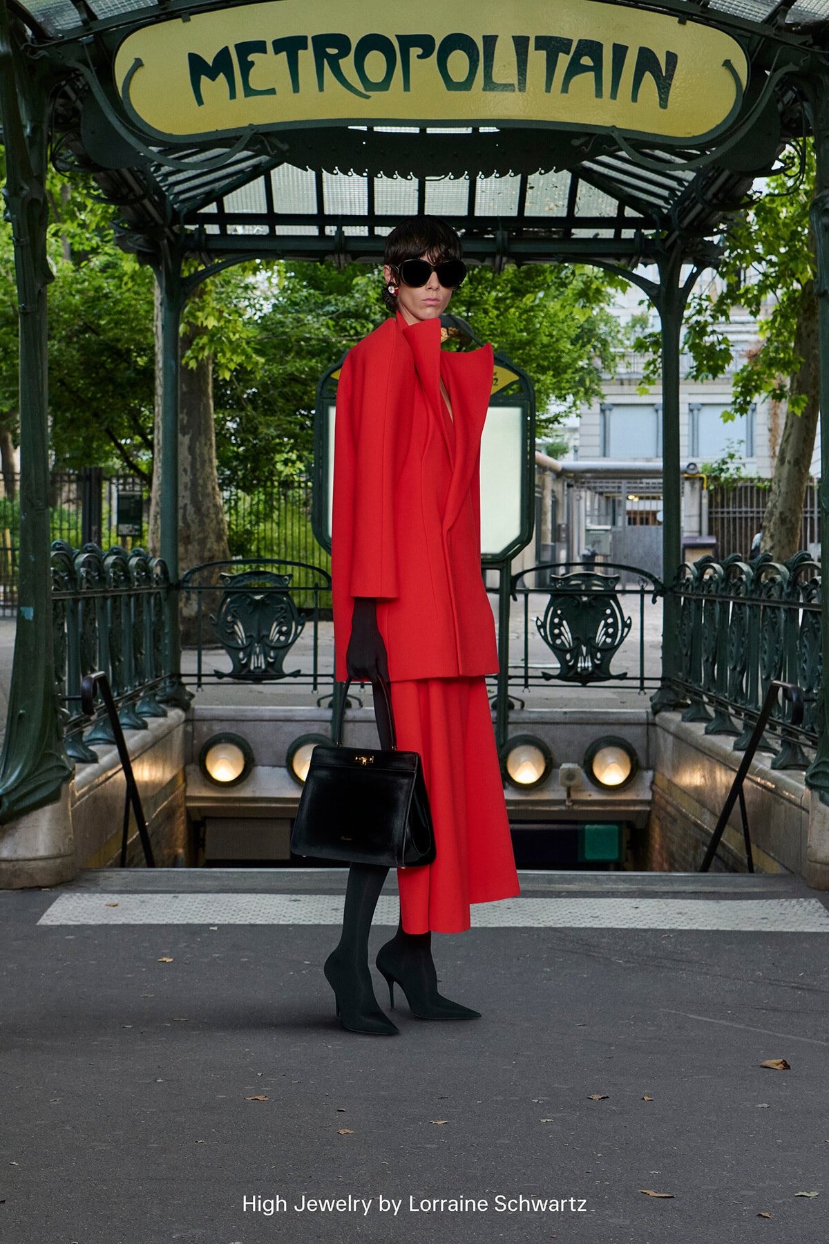 Model in a bright red coat and matching pants with black gloves and handbag posing at a Paris Métropolitain entrance, wearing sunglasses.