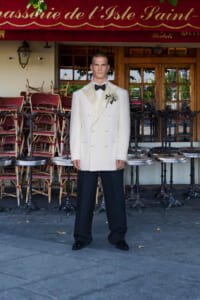 Man in a white double-breasted tuxedo with a black bow tie and boutonniere standing outside a cafe with stacked chairs behind him for ambience.