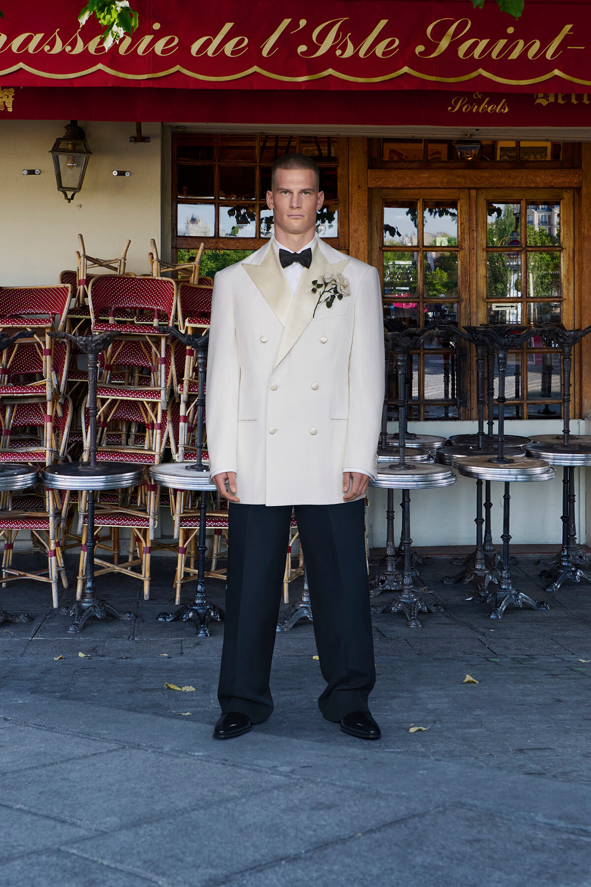 Man in a white double-breasted tuxedo with a black bow tie and boutonniere standing outside a cafe with stacked chairs behind him for ambience.