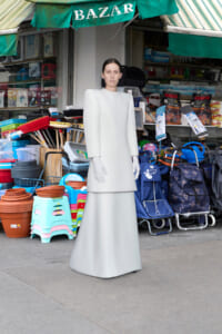 Person wearing a pale gray, sculptural gown with exaggerated shoulders, standing outside a market stall filled with cleaning supplies and goods.