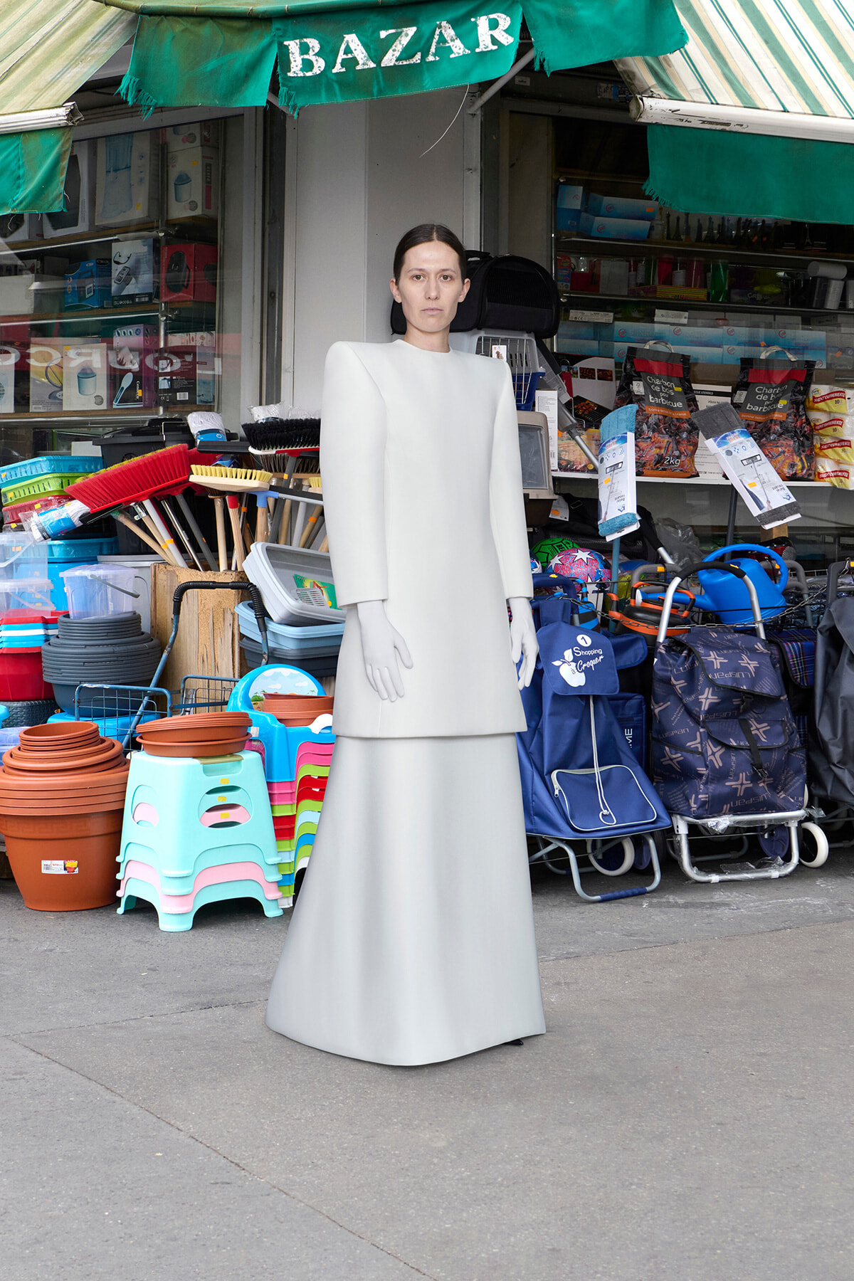 Person wearing a pale gray, sculptural gown with exaggerated shoulders, standing outside a market stall filled with cleaning supplies and goods.
