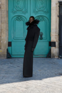 Person wearing a long black coat with a feathered collar stands in front of a turquoise ornate door.