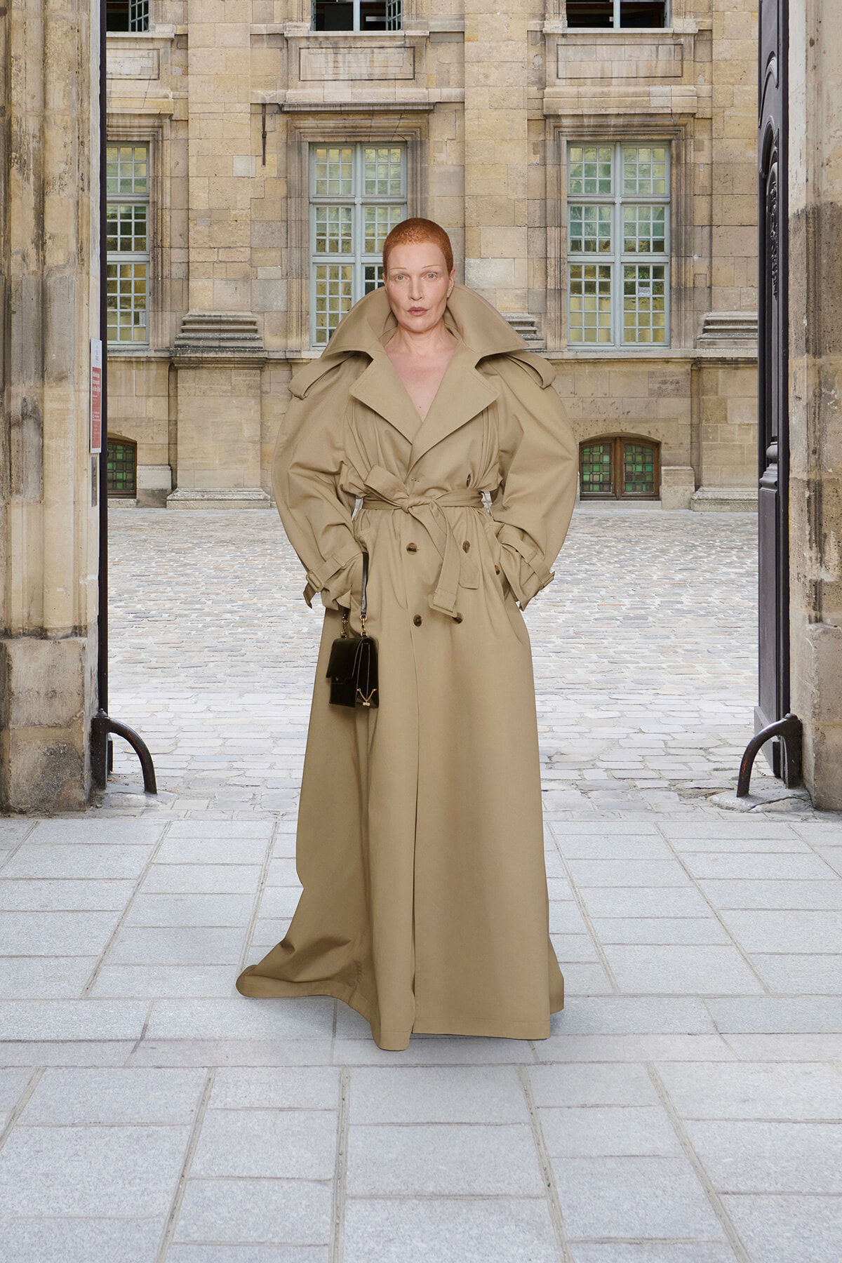 Red-haired person in a long tan trench coat with oversized collar stands in an arched stone courtyard, hands in pockets, holding a small black handbag.