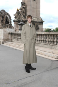 Man in a beige trench coat standing on a stone bridge with ornate statues in the background.