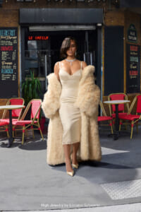 Woman in a cream slip dress and oversized fur coat poses on a city street outside a cafe with red chairs and menus on the wall behind her, wearing statement jewelry.