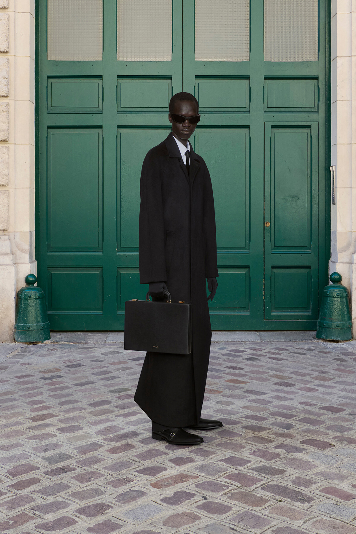 Man in a long black coat and sunglasses holds a black briefcase in front of a green paneled door on a cobblestone street.