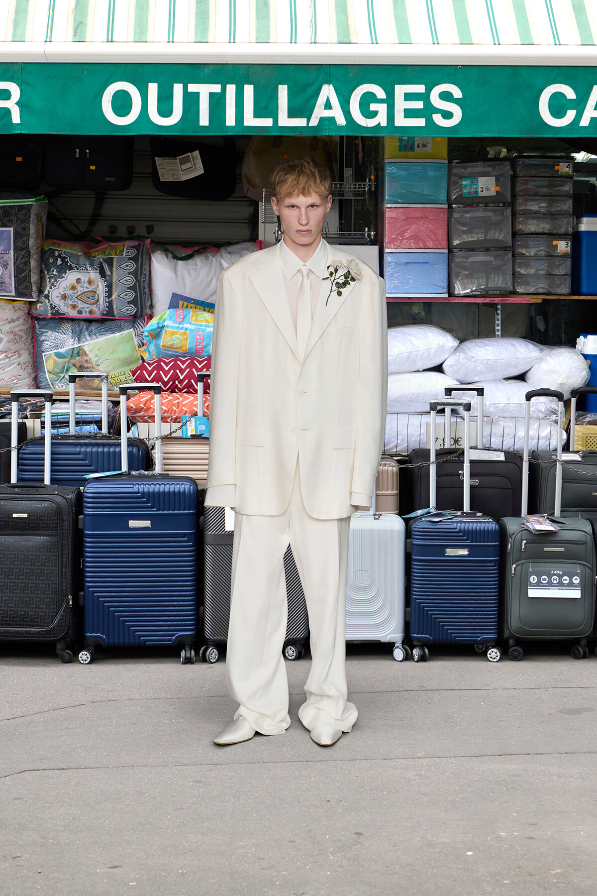 Man in an oversized ivory suit with a boutonniere standing in front of a luggage shop filled with suitcases and pillows outdoors.