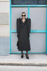 Model in a black polka-dot coat with dramatic shoulders, sunglasses, gloves, and a black handbag against a teal doorway.