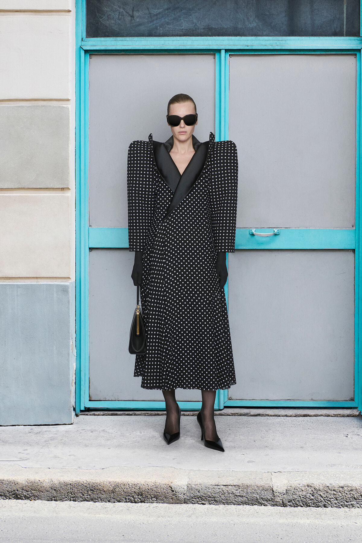 Model in a black polka-dot coat with dramatic shoulders, sunglasses, gloves, and a black handbag against a teal doorway.