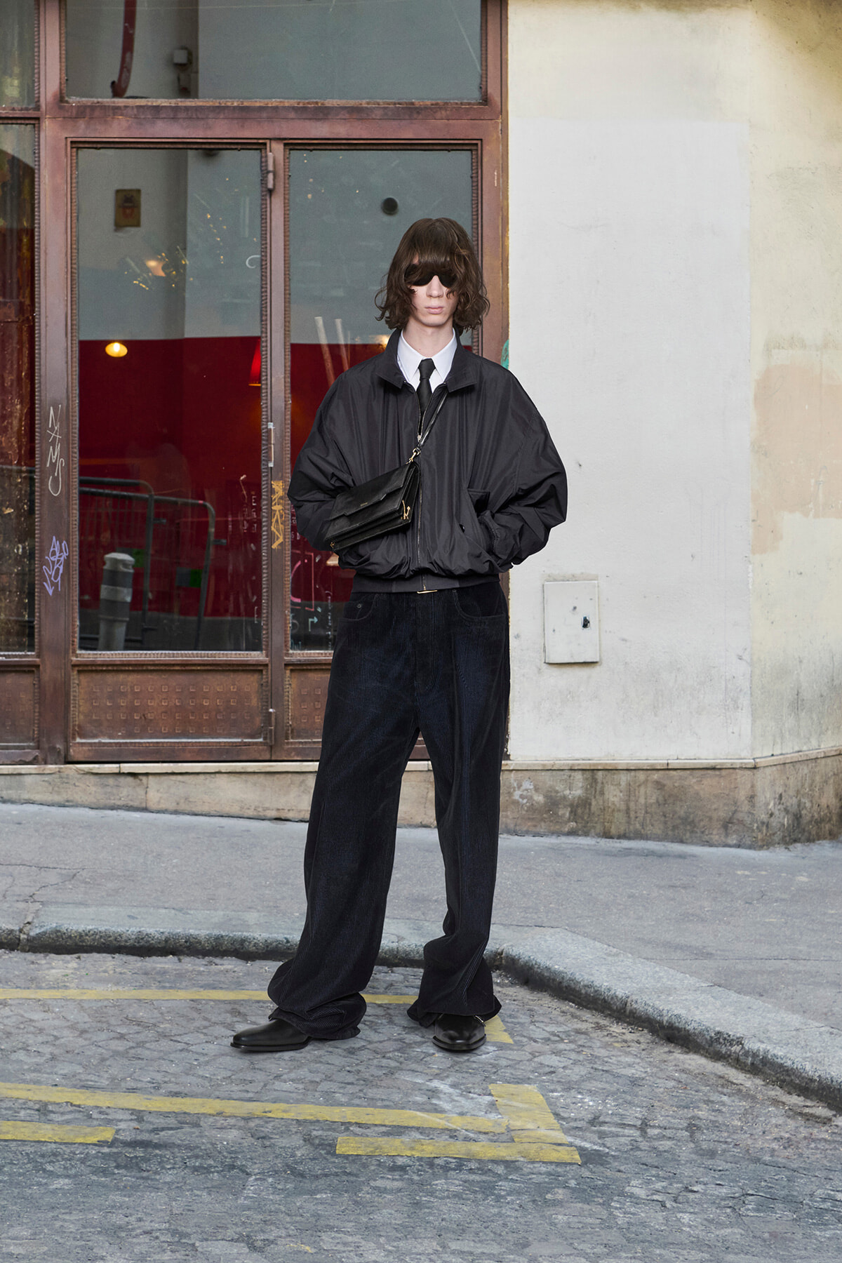 Person standing on a city sidewalk in a black windbreaker, wide-leg dark jeans, and sunglasses, with a crossbody bag in front of a glass storefront.