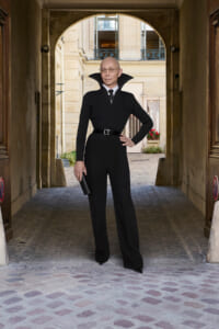 Portrait of a bald man in a black turtleneck and wide-collar coat, with a belt, standing with one hand on his hip under an arched stone passageway.