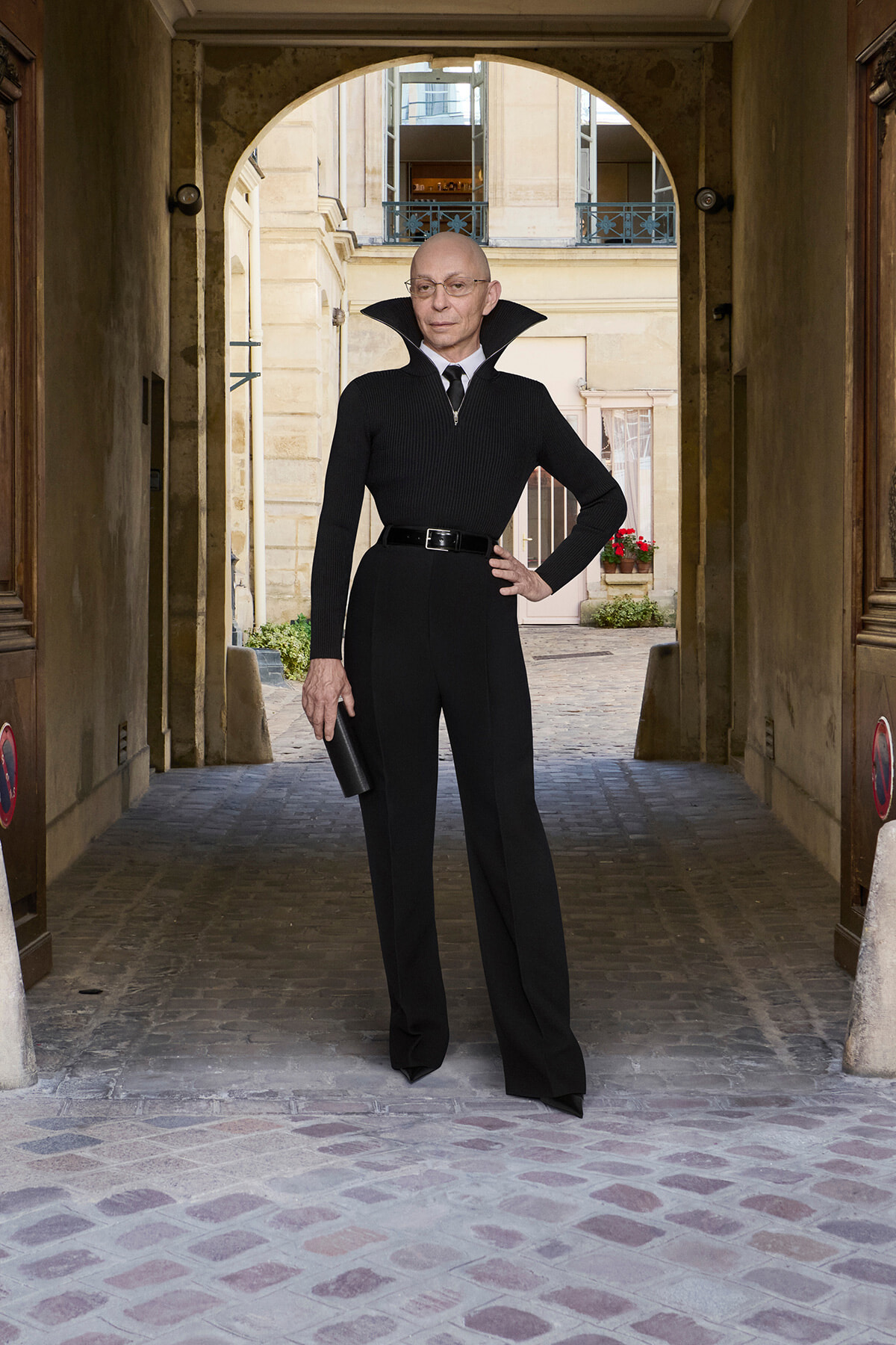 Portrait of a bald man in a black turtleneck and wide-collar coat, with a belt, standing with one hand on his hip under an arched stone passageway.