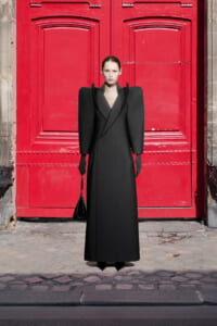Model wearing a dramatic black coat with oversized shoulders and a small black handbag, standing before a bright red door.