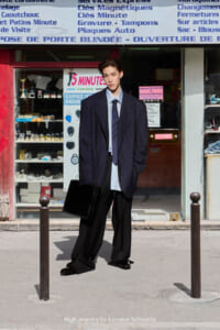 Man in an oversized navy blazer, light shirt and tie, holding a black briefcase on a city sidewalk in front of a shop window.
