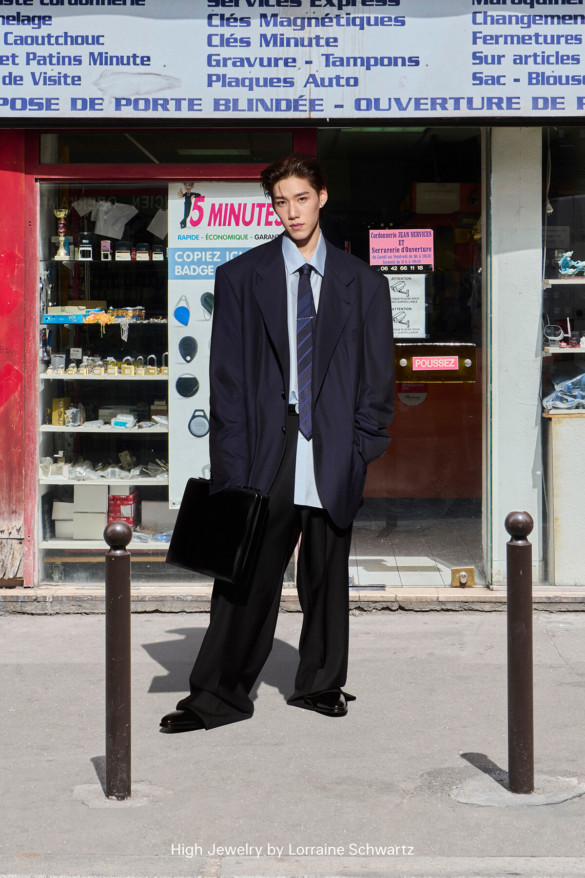 Man in an oversized navy blazer, light shirt and tie, holding a black briefcase on a city sidewalk in front of a shop window.