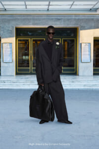 Tall man in an oversized black suit with a bow tie and white boutonniere stands outside a marble building, holding a large black bag and wearing sunglasses.