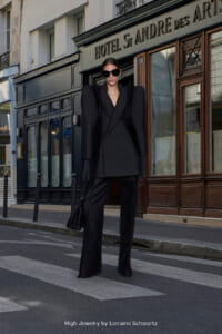 Model in an oversized black pantsuit with dramatic shoulders, black gloves, sunglasses, and a black bag stands on a Paris street in front of Hotel St. André des Arts.