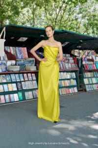 Model in a bright yellow strapless satin gown posing with hands on hips in an outdoor bookstall surrounded by shelves of colorful books. Caption reads 'High Jewelry by Lorraine Schwartz'.