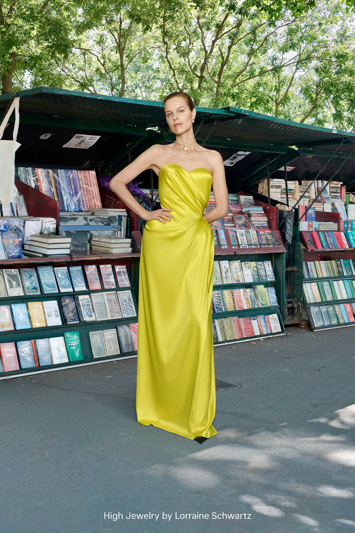Model in a bright yellow strapless satin gown posing with hands on hips in an outdoor bookstall surrounded by shelves of colorful books. Caption reads 'High Jewelry by Lorraine Schwartz'.