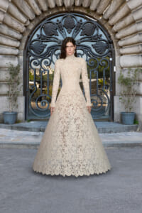 Model wearing a beige lace high-neck wedding gown standing before an ornate arched stone doorway with a wrought-iron gate grew patterning in the metalwork.