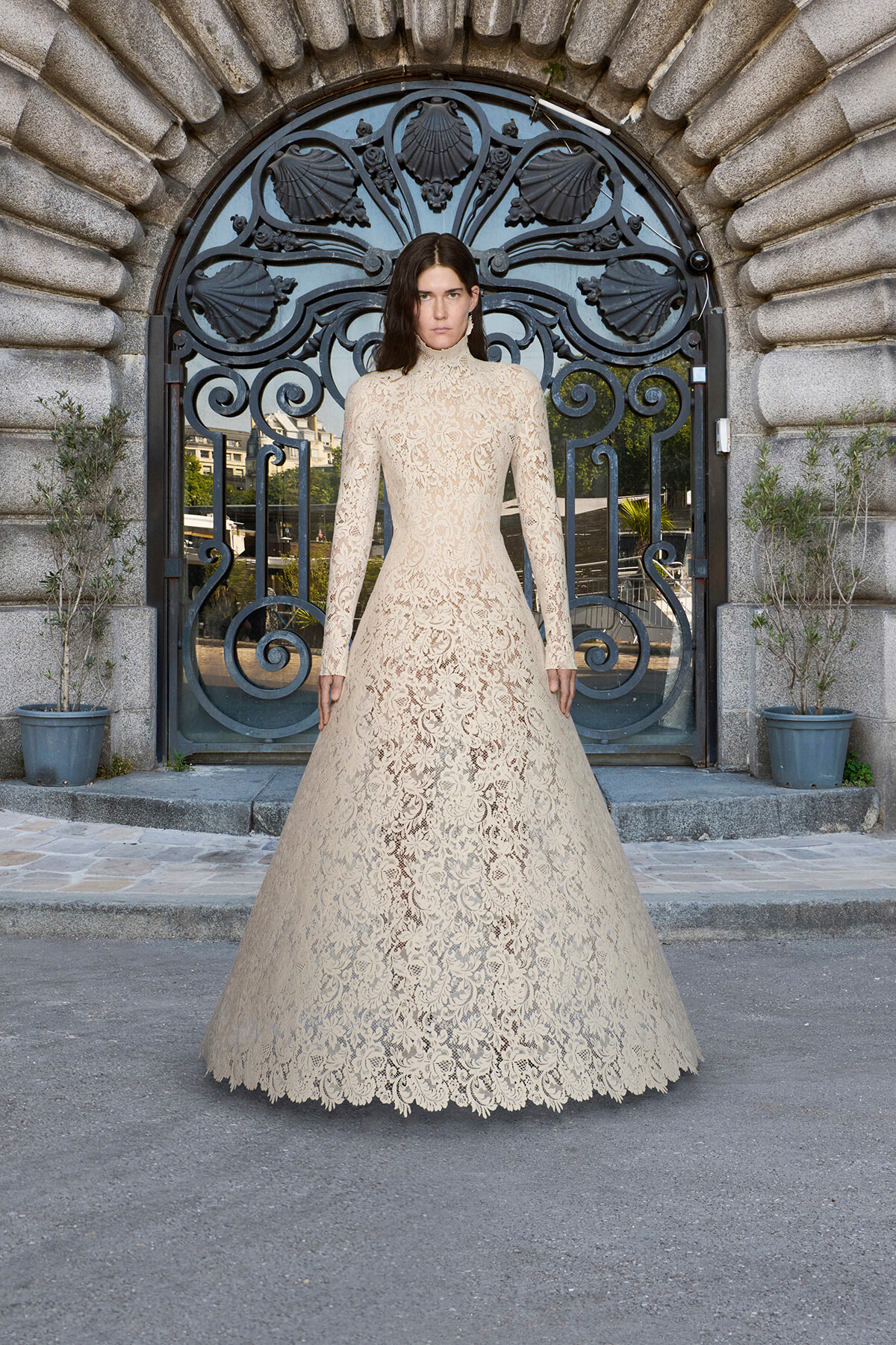 Model wearing a beige lace high-neck wedding gown standing before an ornate arched stone doorway with a wrought-iron gate grew patterning in the metalwork.