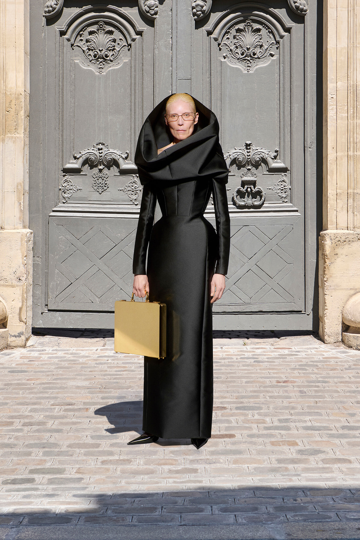 Person in a dramatic black outfit with an oversized collar stands before ornate gray doors, holding a gold briefcase.