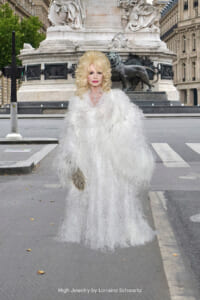 Woman in a voluminous white feather gown and jewelry standing on a city street with a grand stone monument in the background; caption reads 'High Jewelry by Lorraine Schwartz' on the image.