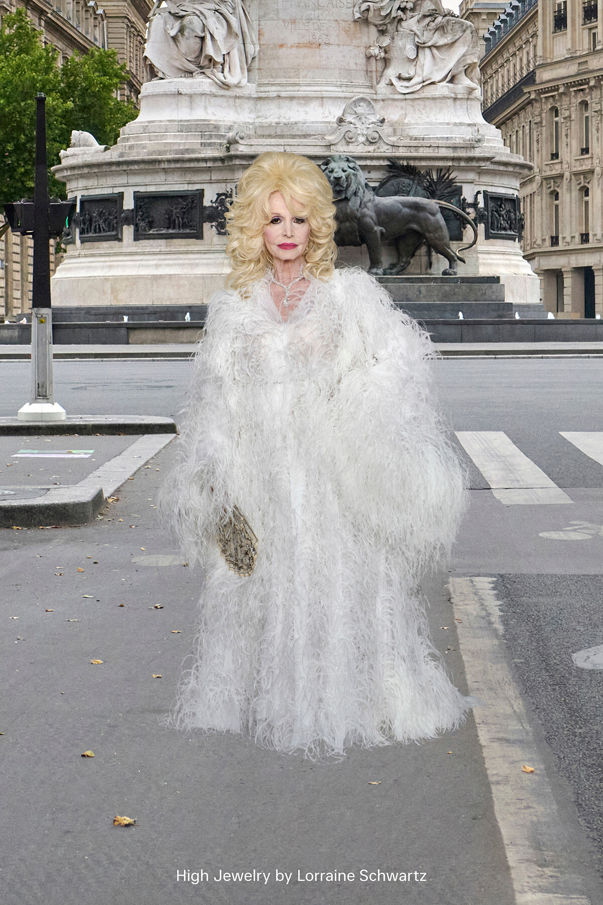Woman in a voluminous white feather gown and jewelry standing on a city street with a grand stone monument in the background; caption reads 'High Jewelry by Lorraine Schwartz' on the image.
