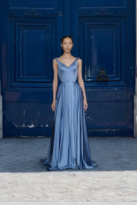 Model wearing a silky light-blue evening gown standing in front of a dark blue ornate door.