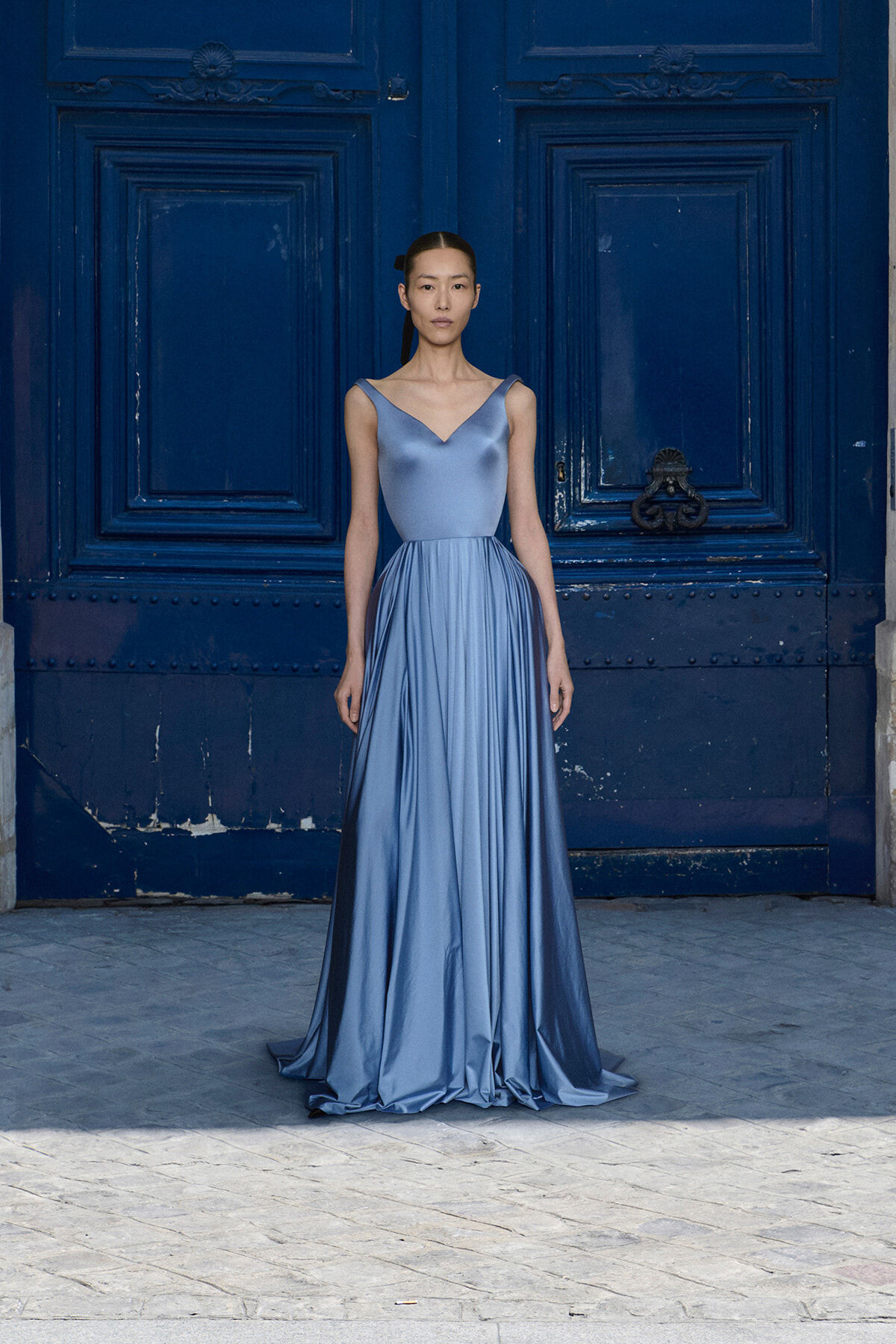 Model wearing a silky light-blue evening gown standing in front of a dark blue ornate door.