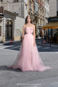 Woman in a pink strapless ball gown with a tiara stands on a sunlit European street, storefronts and a café behind her.