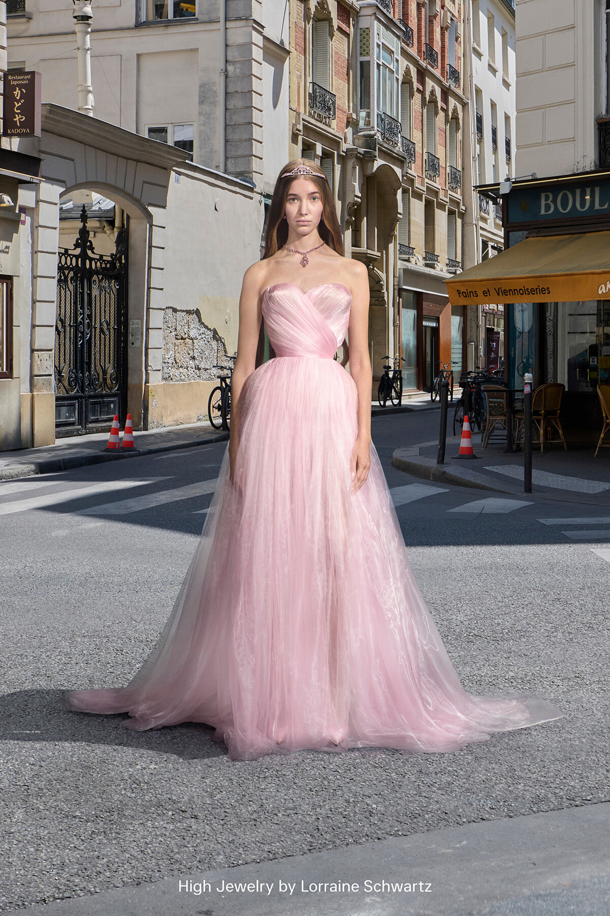 Woman in a pink strapless ball gown with a tiara stands on a sunlit European street, storefronts and a café behind her.