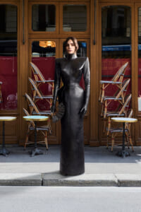 Model in a long, structured black leather dress with high shoulders stands outside a café, clutch in hand.